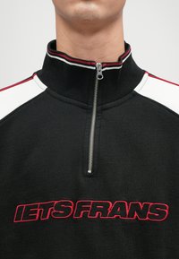 Μαύρο φούτερ με φερμουάρ και υψηλό γιακά, διακοσμημένο με κόκκινες και λευκές λεπτομέρειες στους ώμους και με κεντημένη την ένδειξη "IETS FRANS" σε κόκκινο χρώμα.