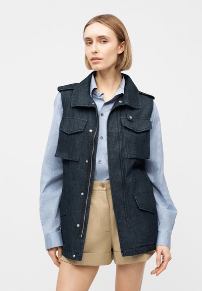Γυναίκα που φοράει ένα σκούρο τζιν γιλέκο πάνω από ένα μπλε ριγέ πουκάμισο με κουμπιά, συνδυασμένο με μπεζ σορτς ψηλόμεσα, σε ουδέτερο φόντο.