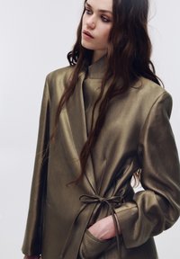 Metallisk olivengrøn blazer med struktureret design, bindebånd i taljen og lange ærmer. Glat tekstur med en subtil glans og sidelommer.