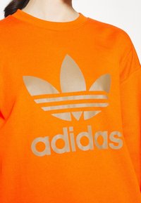 Sudadera de color naranja brillante con un gran logo reflectante beige de Adidas, que incluye tres rayas y un emblema de trefoil en el centro.