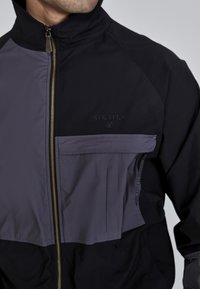 Sort og grå lynlåsjakke med høj krave, der har en frontlomme, guld lynlås og struktureret stof. Brandlogoet er synligt på brystet.