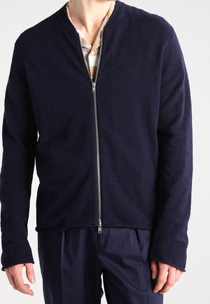 Man draagt een navy blauwe fleecejas met ritssluiting over een gedeeltelijk zichtbare licht gestreepte blouse en donkere broek, staand tegen een effen achtergrond.