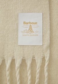 Μπεζ μάλλινο κασκόλ με φριζαριστές άκρες και λευκή ετικέτα με χρυσό κείμενο, απεικονίζοντας έναν φάρο και τις λέξεις "Barbour" και "SOUTH SHIELDS."