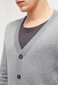 Šedý cardigan s hlubokým výstřihem do V, zdobený žebrovanými detaily a třemi černými knoflíky, přelité přes šedé tričko. Hladká pletená textura.