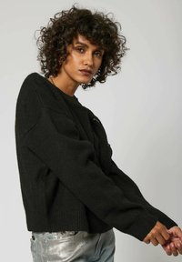 Pull noir en tricot avec une finition texturée, des poignets côtelés et un ourlet court. Présente un petit logo décoratif à l'avant.