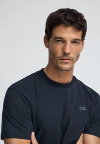 Marineblaue Baumwoll-T-Shirt mit Rundhals, ausgestattet mit einem kleinen rechteckigen Logo-Patch auf der Brust. Kurze Ärmel und eine glatte Textur.