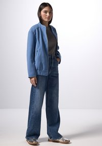 Ljusblå fleecjacka med ståkrage, kombinerad med högmidjade, vida mörkblå jeans och metallicsilverfärgade skor. Slät textur.