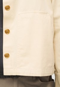 Μπεζ μπουφάν Barbour με καφέ κουμπιά και τσέπη στο στήθος, φορεμένο πάνω από σκούρα ρούχα και συνδυασμένο με καφέ παντελόνια.