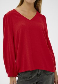 Rode V-hals blouse met loszittende lange mouwen, geweven stof, elastische manchetten en een ontspannen silhouet, gecombineerd met donkere jeans.