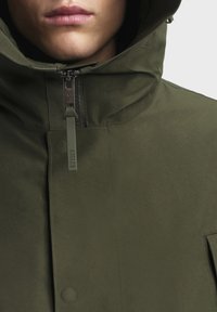 Veste vert olive avec un col haut, fermeture éclair et étiquette de logo. Le matériau semble lisse et résistant à l'eau avec un matériel minimal.