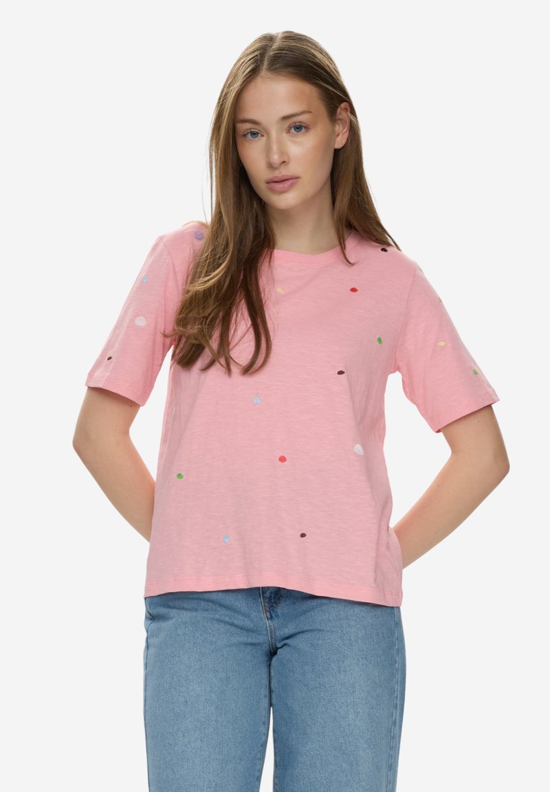 Jeune femme aux longs cheveux portant un t-shirt rose à manches courtes avec de petits points colorés et un jean bleu, debout les mains derrière le dos.
