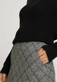 Suéter de punto negro combinado con una falda gris de cintura alta que presenta un sutil patrón de diamantes, con la mano en el bolsillo de la falda.
