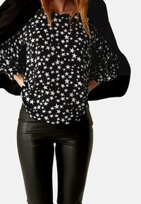 Czarna koszula z długim rękawem z powtarzającym się wzorem białych gwiazd, zestawiona z błyszczącymi czarnymi legginsami, tworząc kontrastujący wygląd.