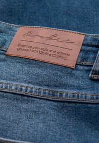 Jean en denim avec une étiquette en cuir marron cousue à la taille, présentant un texte embossé et des coutures distinctes. Le tissu est bleu avec une finition texturée.