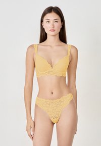 Soutien-gorge en dentelle jaune avec un décolleté plongeant et des bordures festonnées, assorti d'un string en dentelle avec des motifs élaborés.