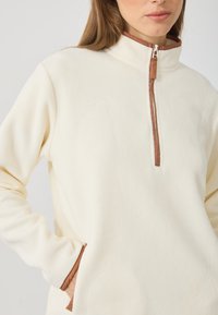 Pulover din fleece de culoare crem cu guler înalt, fermoar la jumătate în față și detalii de accent maro. Are buzunare frontale și o textură netedă.