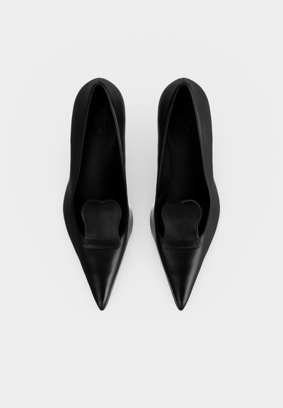 Paire de chaussures en cuir noir à bout pointu sans lacets avec un détail incurvé unique sur le dessus, vues de dessus sur fond blanc.