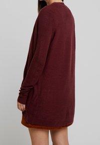 Lång maroon kammad tröja med vertikal ribbad struktur, rund halsringning och lös passform, bärs över en brun kordflugekjol. Mjuk, texturerad tygn.