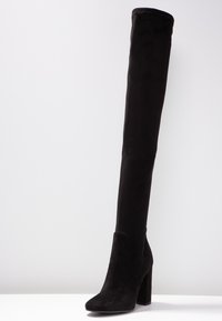 Sort knæhøj støvle lavet af blødt ruskind. Firkantet tå og kraftig hæl. Glat tekstur med minimale detaljer og uden synligt metaludstyr.