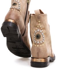 Beżowe zamszowe botki za kostkę mają zamek błyskawiczny, metaliczne akcenty, perłowe zdobienia i czarną okrągłą ozdobę na obcasie.
