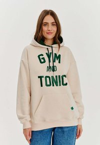 Beige mikina s kapucí a zeleným nápisem "GYM AND TONIC", klokaní kapsa, stahovací šňůrky a žebrované manžety. Vyrobena z měkkého materiálu, volný střih.