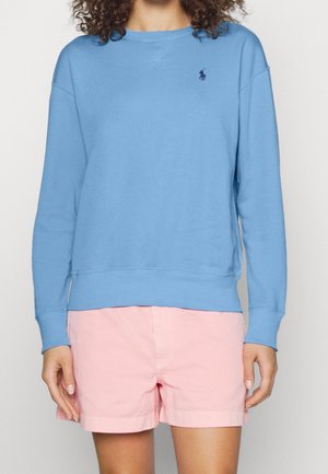 Femme portant un sweat-shirt à manches longues bleu clair avec un petit logo foncé sur la poitrine et un short rose clair, debout devant un fond uni.