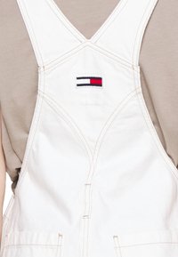 Witte denim tuinbroek gedragen over een beige t-shirt, met een kleine rechthoekige rood-wit-blauwe logo patch op de borst.