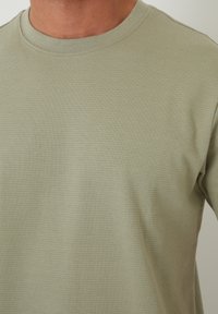 Zbliżenie na osobę w jasnooliwkowej, prążkowanej koszulce z okrągłym dekoltem, ukazującej szyję i górną część klatki piersiowej.