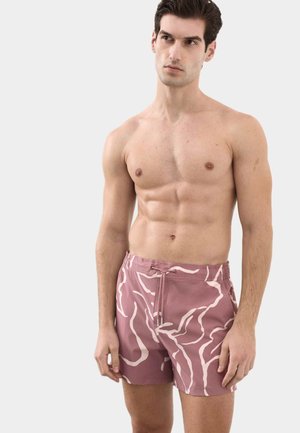 Junger Mann mit kurzem dunklem Haar, der rosa gemusterte Badeshorts trägt und vor einem schlichten hellen Hintergrund steht.