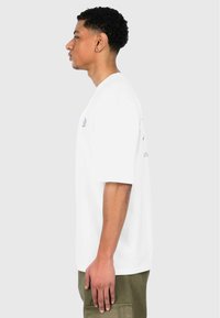 Hvid kortærmet t-shirt med en afslappet pasform, med blå grafiske tryk på fronten og bagsiden, parret med olivengrønne cargobukser.