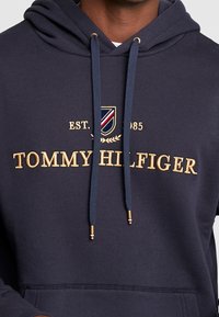 Sudadera azul marino con bordado dorado que dice "TOMMY HILFIGER". Cuenta con capucha ajustable y un logo de escudo con acentos en rojo y blanco.