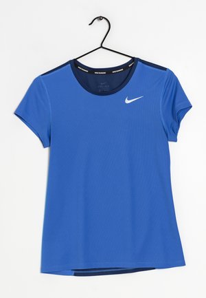 Blå Nike Dri-FIT løbetrøje med korte ærmer og hvid swoosh-logo på brystet, hængende på en sort metalbøjle mod en hvid baggrund.