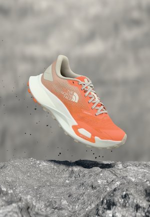 Orangefarbener und beiger Trailrunning-Schuh von The North Face, der über felsigem Gelände schwebt und die profilierte Sohle sowie das seitliche Logo zeigt.