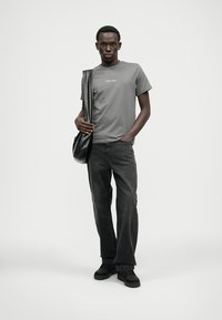 Šedé tričko s logom "REPLAY", spojené s čiernymi džínsami a čiernymi topánkami. Model drží čiernu kabelku cez rameno. Hladká textúra látky.