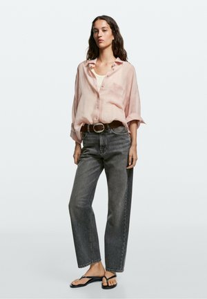 Femme portant une chemise rose ample boutonnée, un haut blanc, un jean gris taille haute avec une ceinture marron, et des tongs noires, debout sur fond blanc.