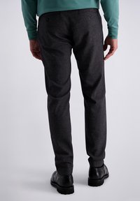 Homme portant un pantalon texturé gris foncé, des chaussures brogues noires et une chemise à manches longues teal, se tenant de dos face à la caméra.