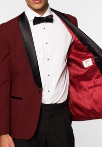 Chaqueta de esmoquin burdeos con solapas de satén negras, un forro interior rojo y un solo botón negro. Combinada con una camisa blanca y pajarita negra.