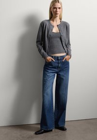 Cardigan grigio lavorato a maglia sopra una canotta grigia a costine, abbinato a jeans a gamba larga in denim blu. Scarpe nere a punta completano il look.