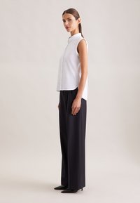 Femme adulte aux cheveux bruns portant une chemise blanche sans manches avec col, un pantalon noir à jambes larges et des talons pointus noirs, debout de profil.
