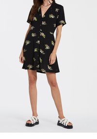 Vestido negro de manga corta con estampados florales en rosa y verde. Cuenta con un cuello y botones en la parte delantera. Sandalias blancas con suela negra.