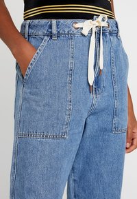 Jasnoniebieskie spodnie dżinsowe z wysokim stanem, bocznymi kieszeniami i sznurkiem do regulacji w brązowych akcentach. Materiał ma teksturowane wykończenie.