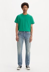 Camiseta de algodón verde, corte relajado, combinada con jeans claros desgastados. El modelo lleva zapatillas negras y blancas. Diseño simple, sin patrones.