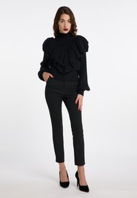 Schwarzer Strickpullover mit geschichteten Rüschen am Halsausschnitt, langen Ärmeln, kombiniert mit enganliegenden schwarzen Hosen und schwarzen High Heels.