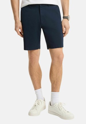 Uomo che indossa pantaloncini blu scuro al ginocchio, calzini bianchi, scarpe da ginnastica bianche, camicia bianca e orologio da polso argento al polso sinistro