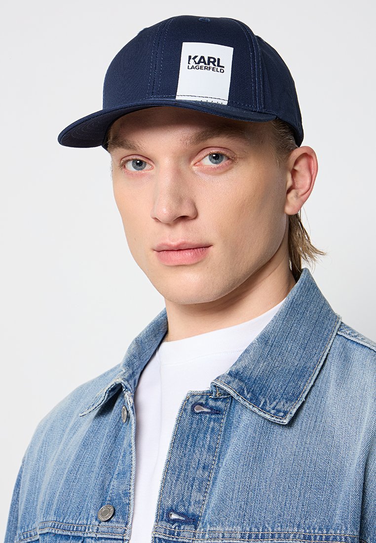 Casquette bleu marine avec une visière plate, présentant une patch rectangulaire blanche avec le logo "KARL LAGERFELD". Portée avec une veste en denim clair.