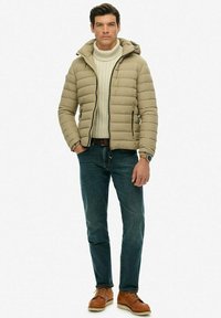Giacca a vento color marrone chiaro, con cappuccio, design trapuntato e due tasche laterali. Indossata sopra un maglione lavorato a maglia color crema, abbinata a jeans blu e scarpe marroni.