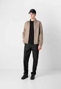 Bežová bunda na zips s elastickými manžetami, čierne tričko, tmavé rifľové nohavice a čierne tenisky. Look dopĺňa čierna čiapka.