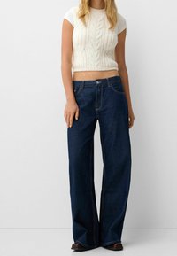 Bílý crop top s pleteným vzorem a krátkými rukávy, který je spárován s tmavě modrými širokými džíny s vysokým pasem a kontrastním šitím.