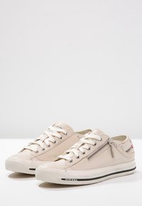 Sneakers basso color beige chiaro con lacci bianchi, materiale in canvas, dettaglio con cerniera laterale, suola in gomma e striscia nera a contrasto sul fondo.