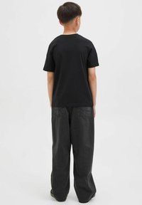 T-shirt en coton noir avec col rond, manches courtes, porté avec un pantalon en denim oversize gris foncé. Design uni visible de dos.